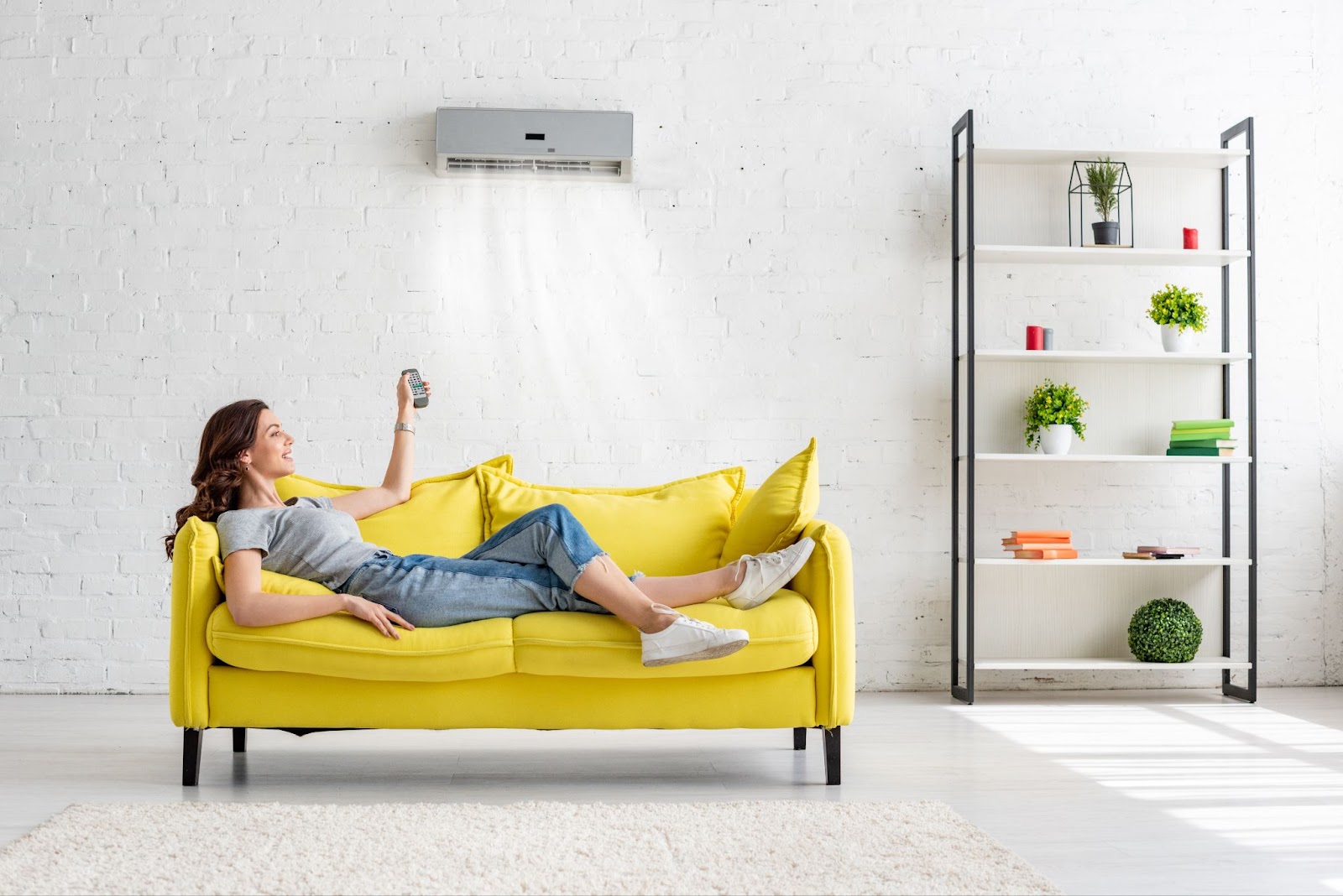 A woman laying on a yellow couch holding a remote control up towards an air conditioning unit on the wall near the ceiling.