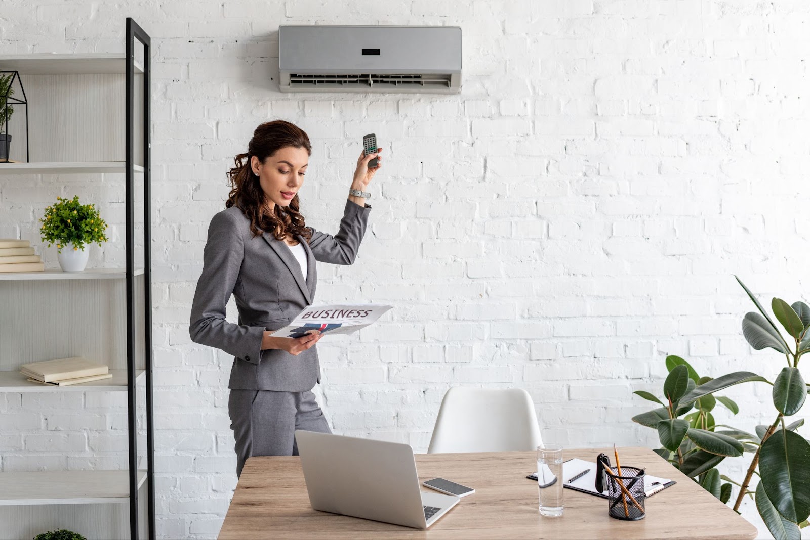 A businesswoman adjusts a ductless AC unit with a remote, engrossed in reading a newspaper. A laptop sits on a table in front of her, with a shelf and plant on her side.