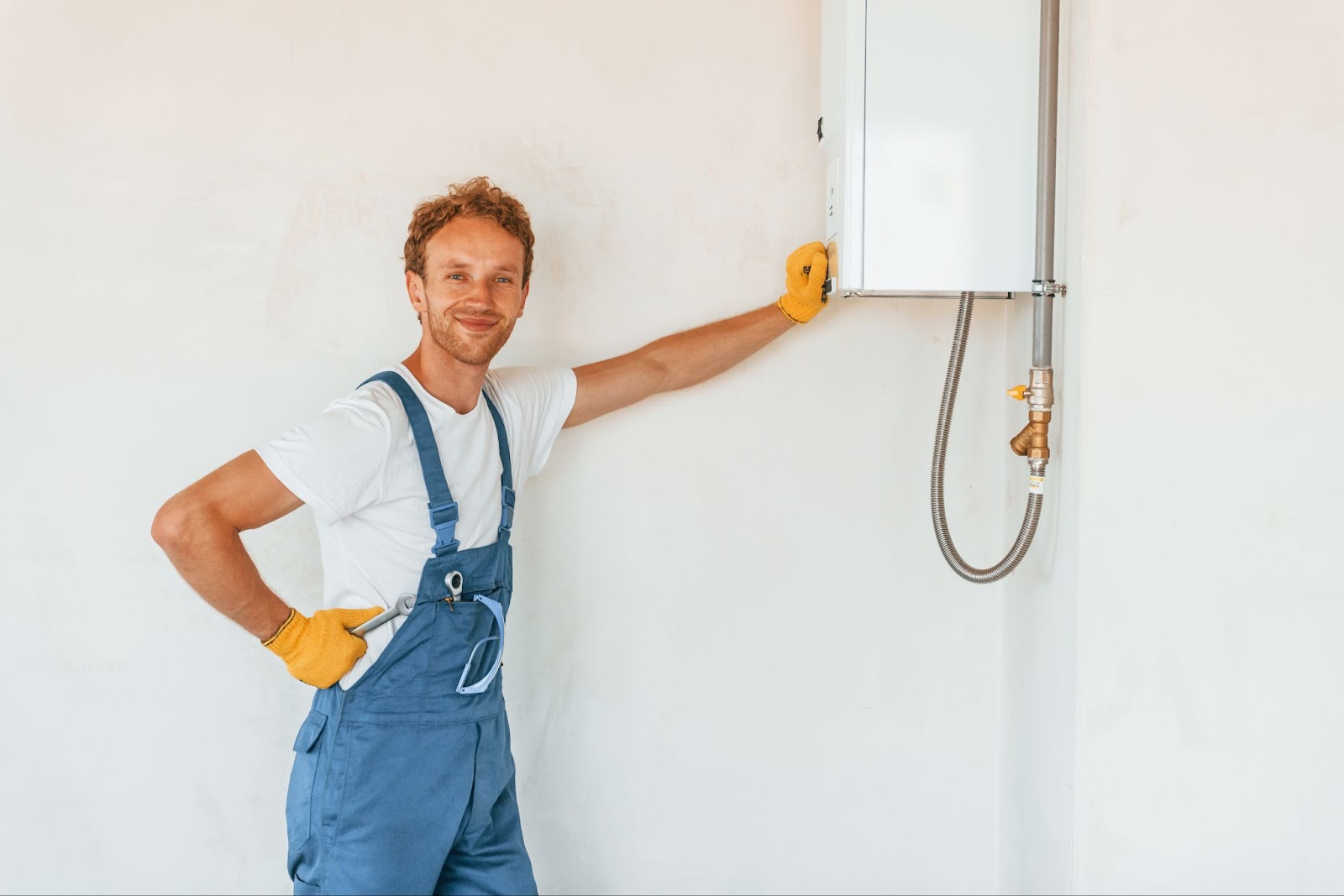 A construction worker wearing overalls and yellow gloves leaning on a newly installed tankless water heater.