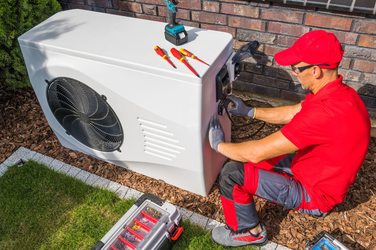 : A technician wearing a red shirt and baseball cap using tools to repair a heat pump outside of someone’s home. 