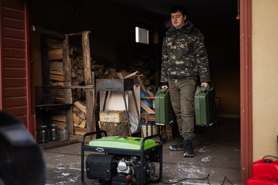 A man wearing camouflage in a shed carrying two canisters of gasoline walking towards an outdoor power generator.